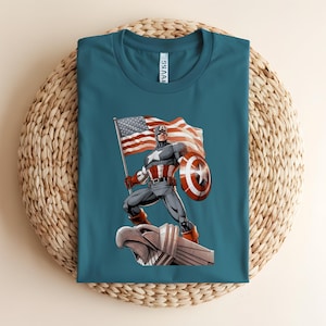 Pode incluir: Camiseta azul-petr&oacute;leo com um gr&aacute;fico do Capit&atilde;o Am&eacute;rica segurando uma bandeira e um escudo americanos. O super-her&oacute;i est&aacute; em p&eacute; sobre uma est&aacute;tua de &aacute;guia. O design &eacute; em estilo de quadrinhos.