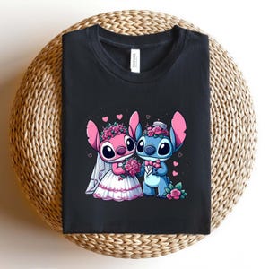 May include: A black crew neck t-shirt featuring a graphic of two cartoon characters dressed for a wedding. One character is pink with a white veil and dress, holding pink roses. The other is blue with a tuxedo and bow tie, also holding roses.