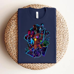 May include: A navy blue t-shirt with a colourful graphic of Avengers characters and logo. The design includes a glowing gauntlet and superhero portraits within geometric shapes. The shirt is displayed on a woven surface.