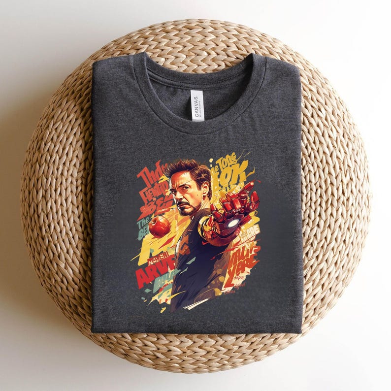 May include: Dark gray t-shirt featuring a colorful graphic of a man with a red and gold armored hand, with text that reads "The Tones OK" and "Marvel Army". The shirt is folded on a woven surface.