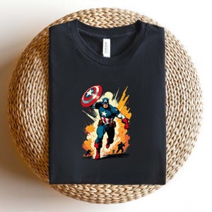 May include: Black t-shirt featuring a Captain America graphic. The illustration shows Captain America in action, throwing his shield with a burst of orange and yellow behind him. The shirt is folded on a woven surface.