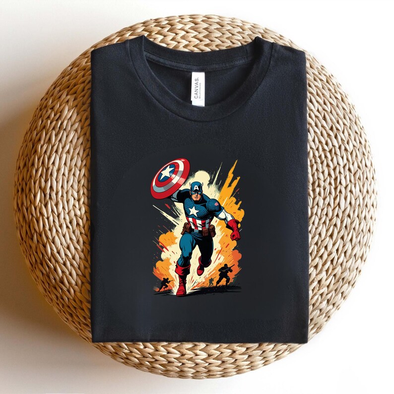 May include: Black t-shirt featuring a Captain America graphic. The illustration shows Captain America in action, throwing his shield with a burst of orange and yellow behind him. The shirt is folded on a woven surface.