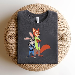 Peut inclure: Un t-shirt gris chin&eacute; fonc&eacute; avec une impression graphique repr&eacute;sentant un lapin de dessin anim&eacute; en uniforme de police bleu et un renard de dessin anim&eacute; portant une chemise vert clair avec une cravate violette. Col rond et &eacute;tiquette blanche.