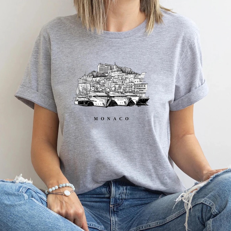 K&ouml;nnte beinhalten: Graues T-Shirt mit einer Schwarz-Wei&szlig;-Illustration der Skyline und Yachten von Monaco. Das Wort "MONACO" ist darunter gedruckt. Das T-Shirt hat hochgekrempelte &Auml;rmel und wird von einer Person getragen.