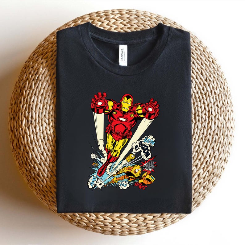 May include: Black t-shirt featuring a comic book style illustration of Iron Man in flight, with a red and yellow color scheme. The graphic includes action lines and a second Iron Man figure below. The shirt is folded on a woven surface.