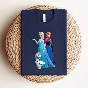 Puede incluir: Camiseta azul marino con un dise&ntilde;o de Elsa, Anna y Olaf de la pel&iacute;cula Frozen. Elsa lleva un vestido azul claro, Anna un vestido azul con una capa rosa y Olaf es un mu&ntilde;eco de nieve blanco. La camiseta est&aacute; doblada sobre una superficie tejida.