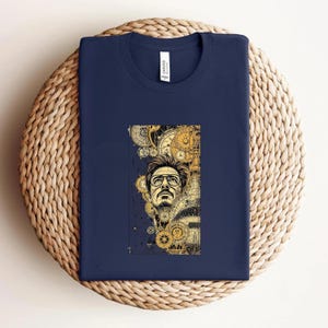 Pode incluir: Camiseta azul marinho com uma ilustra&ccedil;&atilde;o dourada e preta de um homem com &oacute;culos, rodeado por engrenagens e elementos de relojoaria. A camiseta est&aacute; dobrada sobre uma superf&iacute;cie tecida.