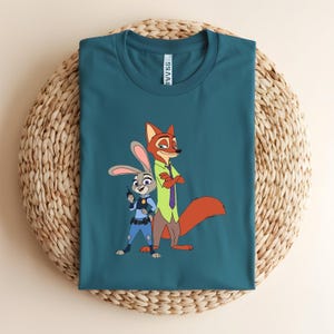 Peut inclure: Un t-shirt bleu sarcelle &agrave; manches courtes avec une impression graphique repr&eacute;sentant un lapin de dessin anim&eacute; en uniforme de police bleu tenant une radio et un renard de dessin anim&eacute; portant une chemise verte et une cravate violette.