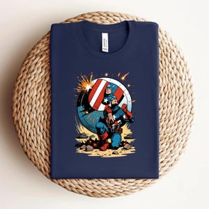 Puede incluir: Camiseta azul marino con una ilustraci&oacute;n de Capit&aacute;n Am&eacute;rica. El gr&aacute;fico muestra al Capit&aacute;n Am&eacute;rica arrodillado con su escudo, con un dise&ntilde;o rojo, blanco y azul. La camiseta est&aacute; doblada sobre una superficie tejida.