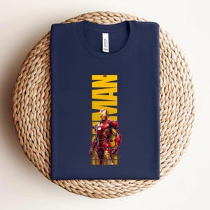 May include: Navy blue t-shirt featuring a graphic of Iron Man in his red and gold armor. The word "MAN" is printed vertically in yellow. The shirt is folded on a woven, circular surface.