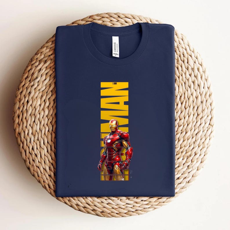 May include: Navy blue t-shirt featuring a graphic of Iron Man in his red and gold armor. The word "MAN" is printed vertically in yellow. The shirt is folded on a woven, circular surface.
