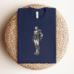 May include: Navy blue t-shirt featuring a detailed illustration of a metallic superhero. The graphic is in shades of silver, gray, and gold. The shirt is folded on a woven, circular surface, with a small white tag visible near the neckline.