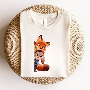May include: A cream-colored t-shirt featuring a graphic print of a cartoon fox and a cartoon rabbit peeking out from behind a vertical edge. The fox has orange fur and large ears, while the rabbit wears a blue uniform with a badge.