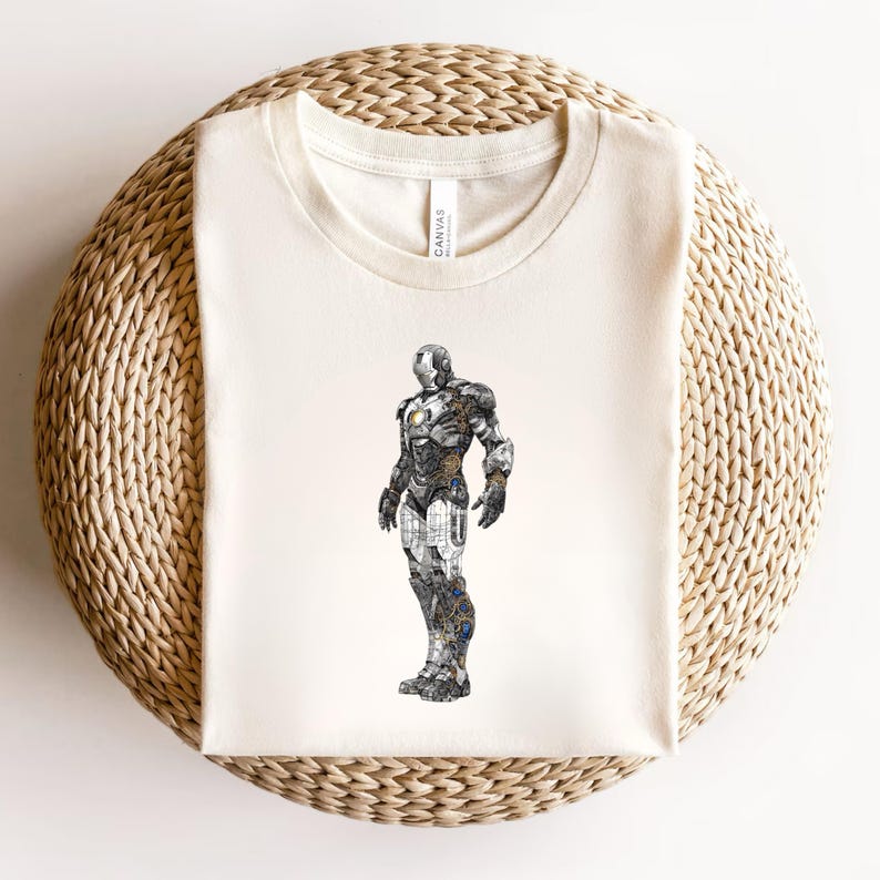 May include: Cream-colored t-shirt featuring a detailed illustration of the Iron Man suit. The artwork showcases the suit's metallic design with intricate mechanical details in shades of silver, gold, and blue. The shirt is laid flat on a woven surface.