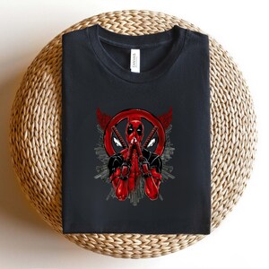 K&ouml;nnte beinhalten: Schwarzes T-Shirt mit einer rot-schwarzen Deadpool-Grafik. Das Design zeigt Deadpools Maske, Schwerter und ein kreisf&ouml;rmiges Emblem. Das T-Shirt liegt gefaltet auf einer gewebten Oberfl&auml;che.