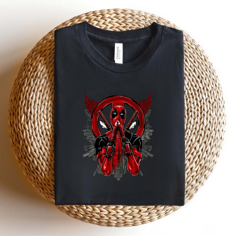 K&ouml;nnte beinhalten: Schwarzes T-Shirt mit einer rot-schwarzen Deadpool-Grafik. Das Design zeigt Deadpools Maske, Schwerter und ein kreisf&ouml;rmiges Emblem. Das T-Shirt liegt gefaltet auf einer gewebten Oberfl&auml;che.