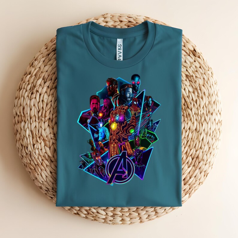 May include: Teal t-shirt with a colourful graphic of Avengers characters and logo. The design includes a glowing gauntlet and geometric shapes, creating a dynamic composition. The shirt is folded on a woven surface.