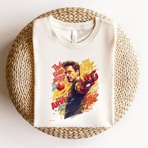 May include: Off-white t-shirt featuring a colorful graphic of a man with a red and gold mechanical arm, pointing forward. The design includes text in red, yellow, and white, with the word "ARVE" visible. The shirt is folded on a woven surface.