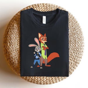 Peut inclure: Un t-shirt noir avec une impression graphique repr&eacute;sentant un lapin de dessin anim&eacute; en uniforme de police bleu et un renard de dessin anim&eacute; en chemise verte et cravate violette. L'&eacute;tiquette blanche au col porte le nom de marque Canvas.