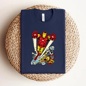 May include: Navy blue t-shirt featuring a vibrant Iron Man graphic in red, yellow, and gold. The illustration shows Iron Man in flight, with comic book sound effects. The shirt is displayed on a woven surface.