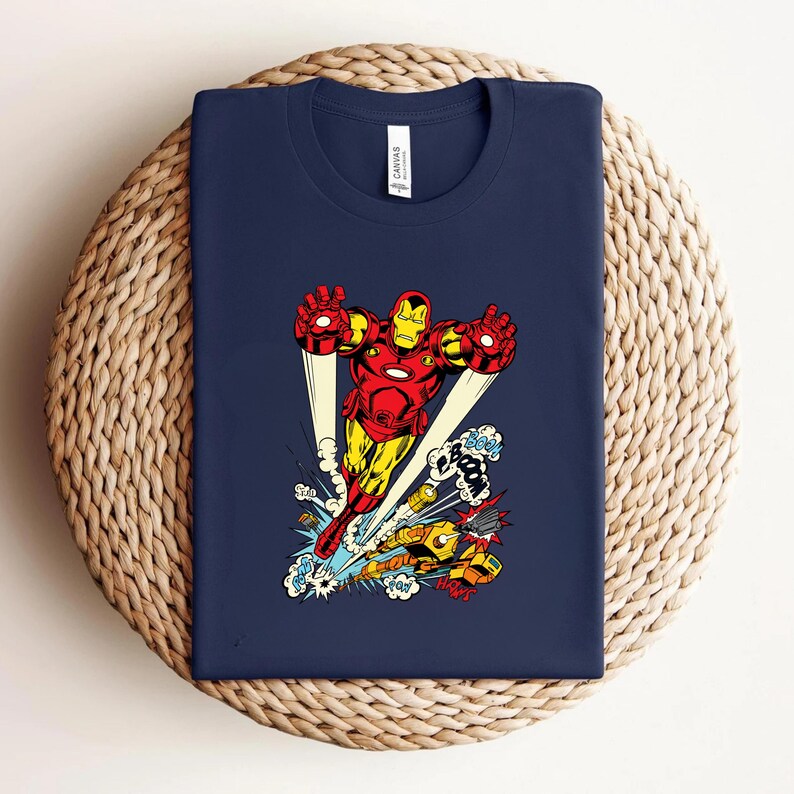 May include: Navy blue t-shirt featuring a vibrant Iron Man graphic in red, yellow, and gold. The illustration shows Iron Man in flight, with comic book sound effects. The shirt is displayed on a woven surface.