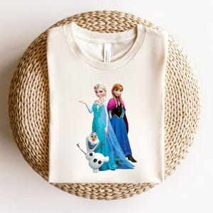 Puede incluir: Camiseta de color crema con un gr&aacute;fico de Elsa, Anna y Olaf de la pel&iacute;cula Frozen. Elsa lleva un vestido azul, Anna un vestido azul y una capa rosa, y Olaf es un mu&ntilde;eco de nieve blanco. La camiseta est&aacute; sobre una superficie circular tejida.