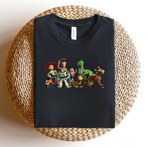 May include: Black t-shirt featuring a colorful graphic of characters from the animated movie Toy Story. The design includes Woody, Buzz Lightyear, Jessie, Rex, and other characters. The shirt is folded on a woven surface.
