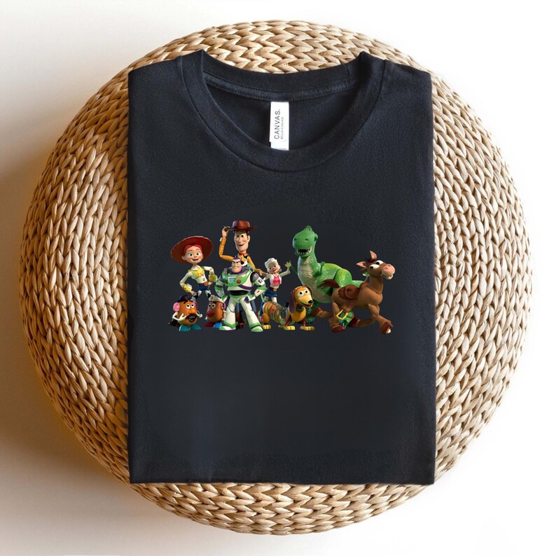 May include: Black t-shirt featuring a colorful graphic of characters from the animated movie Toy Story. The design includes Woody, Buzz Lightyear, Jessie, Rex, and other characters. The shirt is folded on a woven surface.