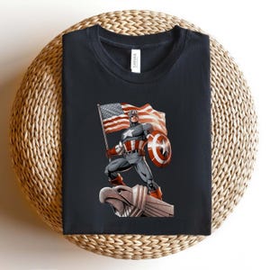 Pode incluir: Camiseta preta com um gr&aacute;fico do Capit&atilde;o Am&eacute;rica segurando uma bandeira americana e um escudo. A ilustra&ccedil;&atilde;o mostra o Capit&atilde;o Am&eacute;rica em uma &aacute;guia de pedra. A camisa tem gola redonda e &eacute; feita de um material macio.