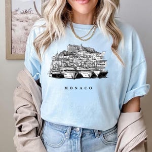 K&ouml;nnte beinhalten: Hellblaues T-Shirt mit einer Schwarz-Wei&szlig;-Illustration der Skyline von Monaco und Yachten. Das Wort "MONACO" ist in Schwarz darunter gedruckt. Das T-Shirt besteht aus einem weichen Material.