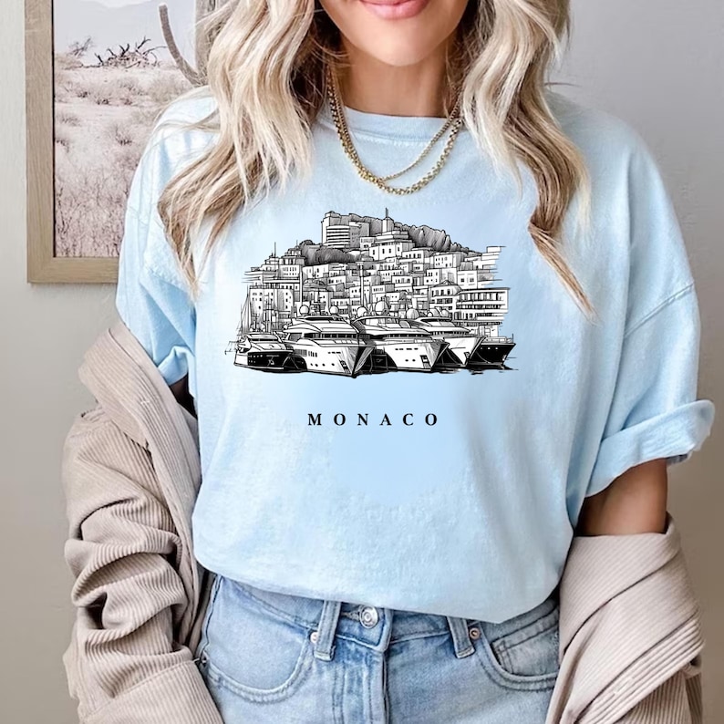 K&ouml;nnte beinhalten: Hellblaues T-Shirt mit einer Schwarz-Wei&szlig;-Illustration der Skyline von Monaco und Yachten. Das Wort "MONACO" ist in Schwarz darunter gedruckt. Das T-Shirt besteht aus einem weichen Material.