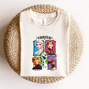 May include: Cream-colored t-shirt featuring the word "FROZEN" above a graphic of Elsa, Anna, Olaf, Kristoff, and Sven. The characters are in colorful rectangular frames. The shirt is folded on a woven, circular surface.