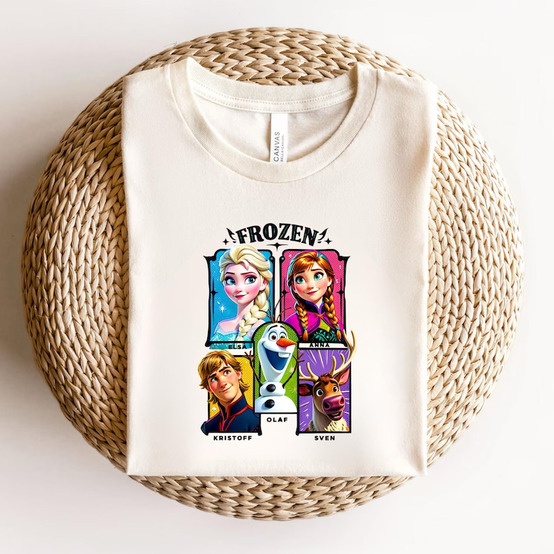 May include: Cream-colored t-shirt featuring the word "FROZEN" above a graphic of Elsa, Anna, Olaf, Kristoff, and Sven. The characters are in colorful rectangular frames. The shirt is folded on a woven, circular surface.
