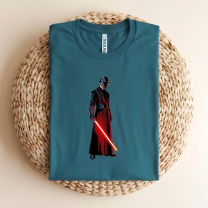 May include: Teal-colored t-shirt featuring a graphic of a character in a black and red outfit, holding a red lightsaber. The character is wearing a helmet and a long robe. The shirt is displayed on a woven, circular mat.