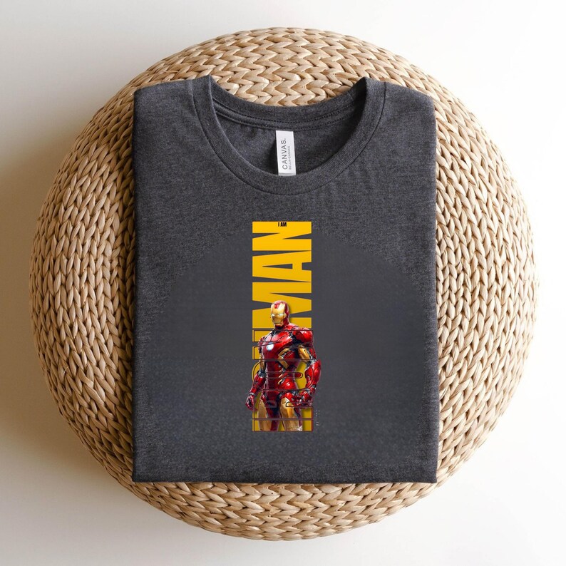 May include: A dark gray t-shirt featuring a graphic of Iron Man in his red and gold armor. The word "IRON MAN" is printed vertically in yellow. The shirt is made of a soft material and is displayed on a woven surface.