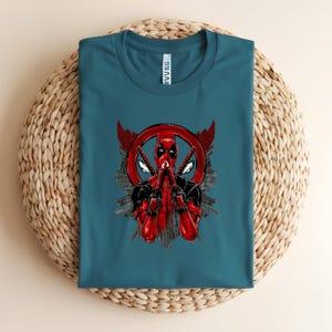 K&ouml;nnte beinhalten: T&uuml;rkisfarbenes T-Shirt mit einem Deadpool-Motiv. Das Design zeigt Deadpool in einem rot-schwarzen Anzug, der zwei Katanas h&auml;lt, vor einem roten Kreis mit Fl&uuml;geln und einer Stadtlandschaft. Das T-Shirt ist auf einer gewebten, kreisf&ouml;rmigen Oberfl&auml;che gefaltet.