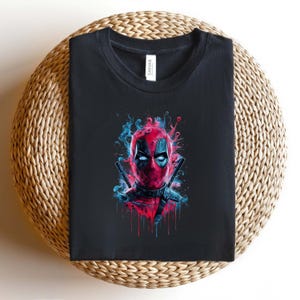 May include: Black t-shirt featuring a colorful graphic of Deadpool's face. The design includes red, blue, and black elements, with two katanas crossed behind the character's head. The shirt is folded on a woven surface.