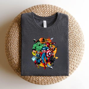 May include: A dark heather gray crew neck t-shirt featuring a colorful graphic print of various comic book superheroes, including Hulk, Thor, Captain America, and Iron Man, arranged in a circular composition with a planet in the background.