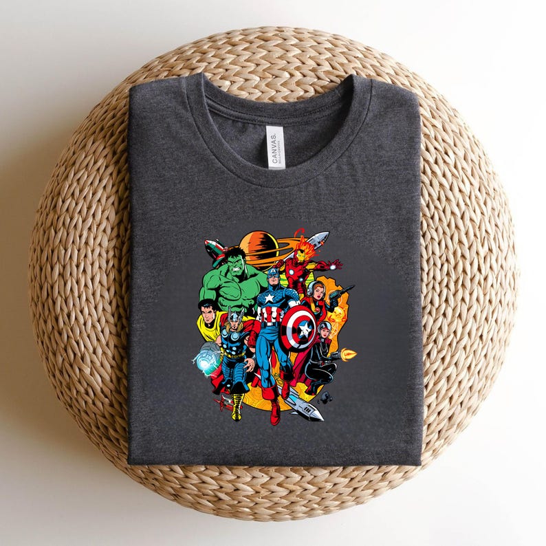 May include: A dark heather gray crew neck t-shirt featuring a colorful graphic print of various comic book superheroes, including Hulk, Thor, Captain America, and Iron Man, arranged in a circular composition with a planet in the background.