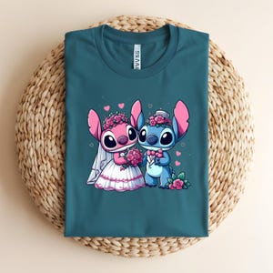 May include: A teal crew neck t-shirt featuring a graphic of two cartoon characters dressed for a wedding. One character is pink with a white veil and flower crown, holding a bouquet. The other is blue with a top hat and bowtie, both with pink hearts.