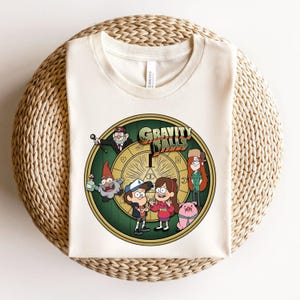 May include: A cream-colored crew neck t-shirt featuring a circular graphic print of the Gravity Falls cast, including Dipper, Mabel, Grunkle Stan, Wendy, a gnome, and Waddles the pig, set against a yellow and green background with the show title.