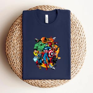 May include: A navy blue crew neck t-shirt featuring a colorful graphic print of various comic book superheroes including Hulk, Thor, Captain America, and Iron Man arranged in a circular composition against a yellow and orange background.