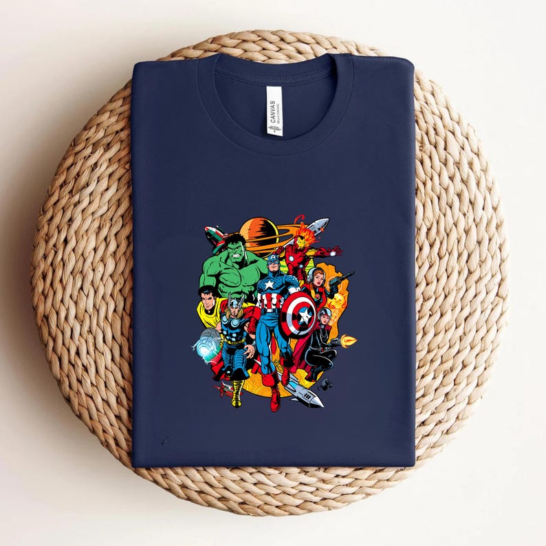 May include: A navy blue crew neck t-shirt featuring a colorful graphic print of various comic book superheroes including Hulk, Thor, Captain America, and Iron Man arranged in a circular composition against a yellow and orange background.