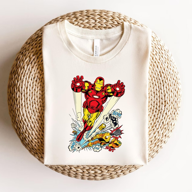May include: Off-white t-shirt featuring a comic book illustration of Iron Man in flight, rendered in red, yellow, and black. The design includes action bubbles with sound effects. The shirt is displayed on a woven, circular surface.
