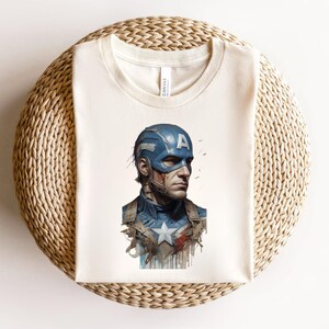 May include: Cream-colored t-shirt featuring a Captain America illustration. The graphic depicts Captain America in his blue helmet with the letter "A", and a blue and white uniform. The shirt is folded on a woven, beige surface.