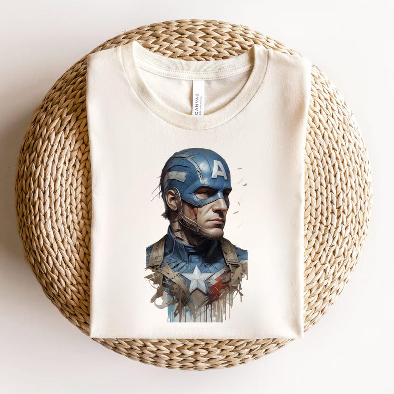 May include: Cream-colored t-shirt featuring a Captain America illustration. The graphic depicts Captain America in his blue helmet with the letter "A", and a blue and white uniform. The shirt is folded on a woven, beige surface.