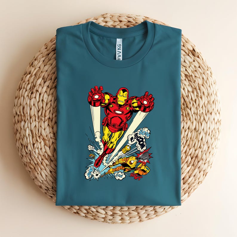 May include: Teal t-shirt featuring a vibrant graphic of Iron Man in flight, with a yellow and red color scheme. The design includes comic book style sound effects and a second Iron Man figure. The shirt is displayed on a woven surface.