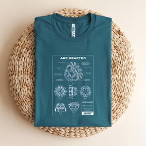Puede incluir: Camiseta de color turquesa con un esquema detallado de un Reactor Arc, con el texto "ARC REACTOR" en la parte superior. El dise&ntilde;o incluye diagramas t&eacute;cnicos y etiquetas. La camiseta tambi&eacute;n tiene el logotipo de Stark Industries.