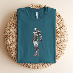 May include: Teal-colored t-shirt featuring a detailed illustration of a metallic superhero. The superhero design incorporates silver, gray, and gold tones, creating a striking contrast against the shirt's background. The shirt is laid on a woven, beige surface.