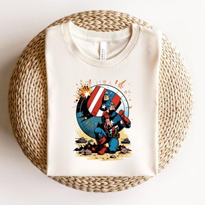 Puede incluir: Camiseta de color crema con un gr&aacute;fico del Capit&aacute;n Am&eacute;rica. El dise&ntilde;o muestra al Capit&aacute;n Am&eacute;rica arrodillado con su escudo, un escudo rojo, blanco y azul, y explosiones. La camiseta est&aacute; doblada sobre una superficie circular tejida.